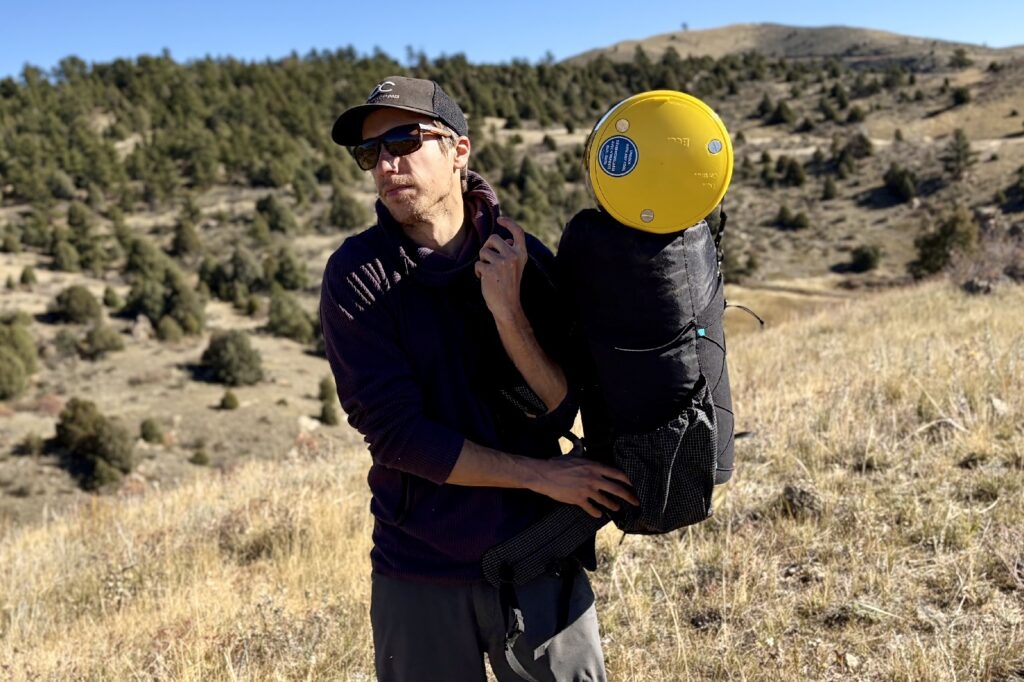 a hiker shoulder his backpack with a yellow bear canister strapped on top