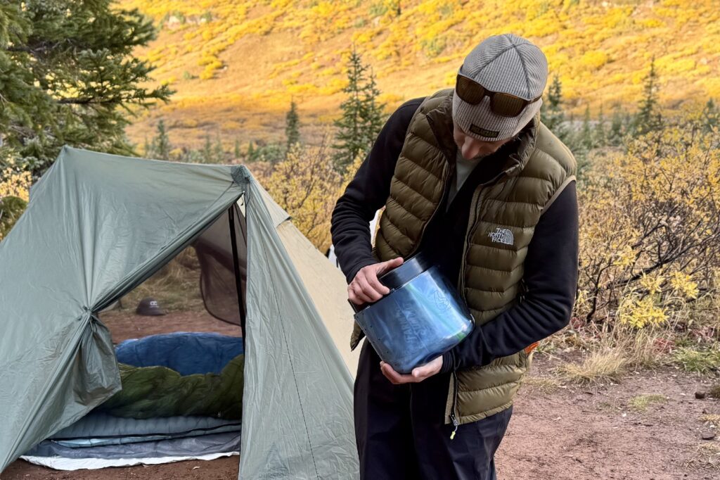 a hiker tries to open a blue bear canister with a tent in the background