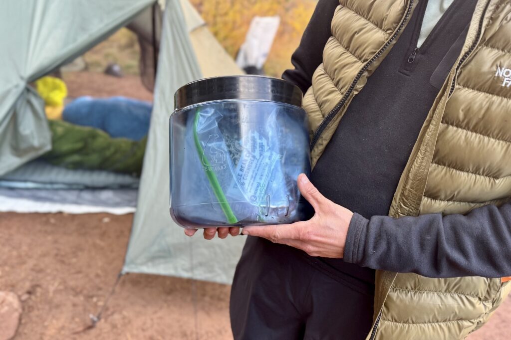 a hiker holds a bear canister full of food with a tent in the background