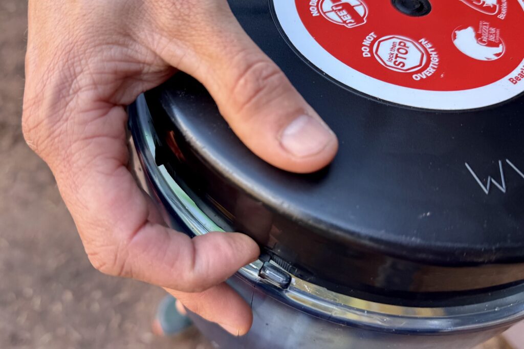 a close up of a hand pushing a black lid of a bear canister to pass the locking tabs
