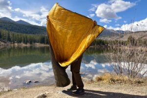 a hiker holds up a grey quilt to show the yellow inside to the camera. a lake a mountains are in the background.