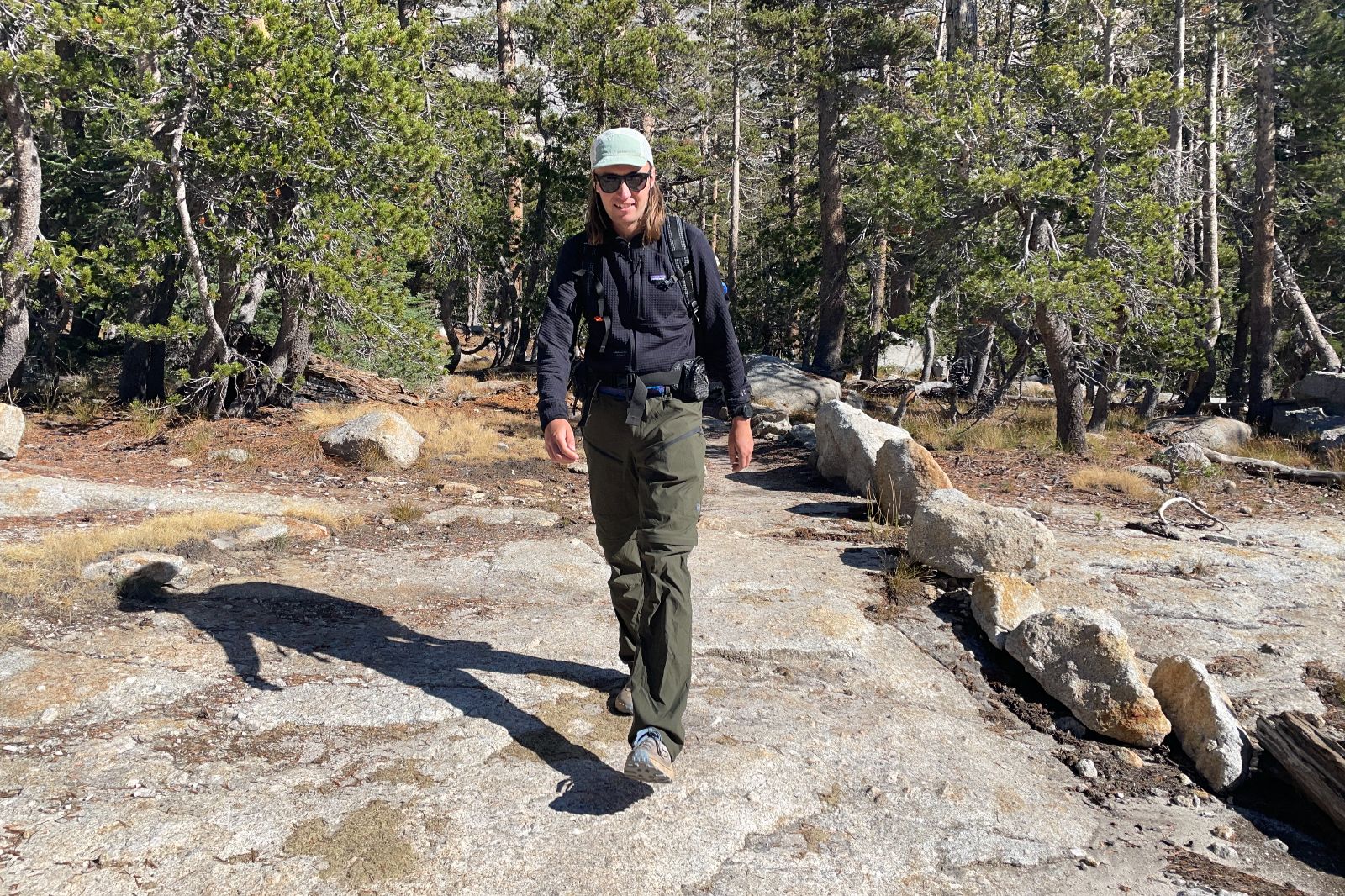 A man walks down a rock-lined trail wearing green pants and a black fleece.