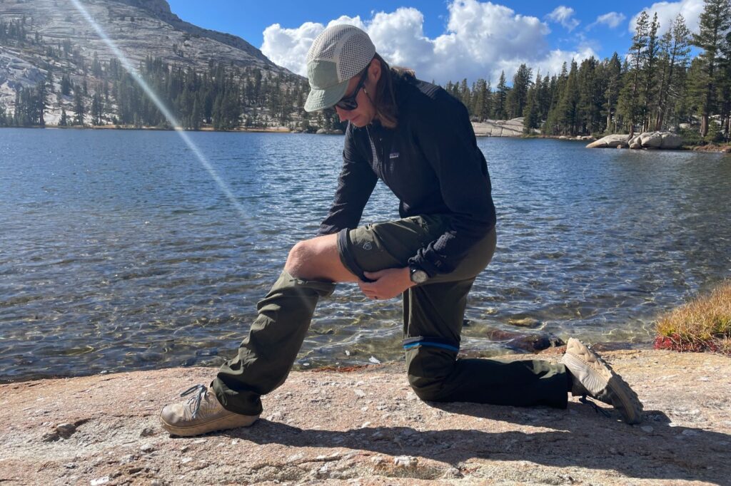 A man converts his convertible pants into shorts alongside a lake.
