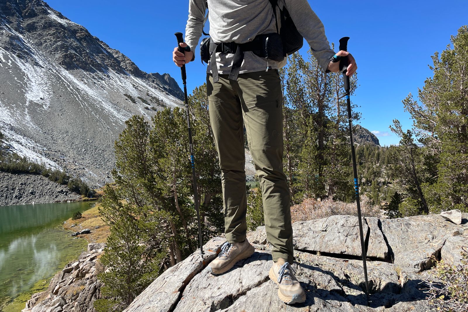 A man stands on a rocky wearing green hiking ants above an alpine lake.