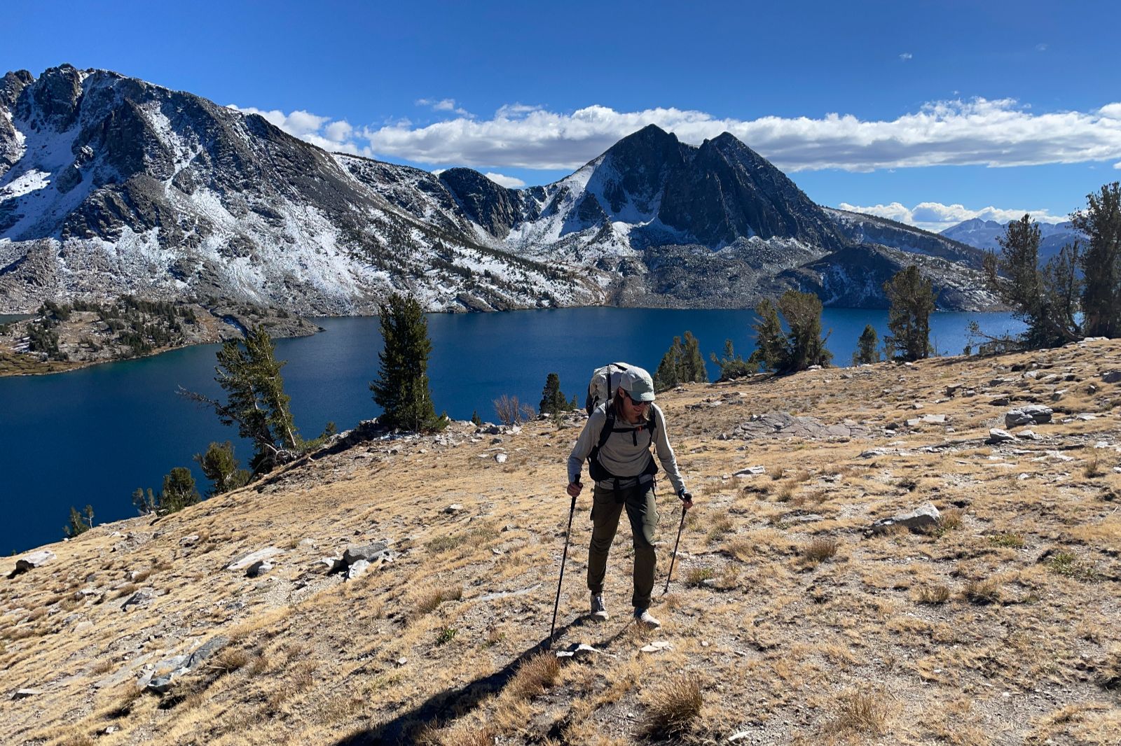 A man hikes above an alpine lake with snowy mountains in the background.