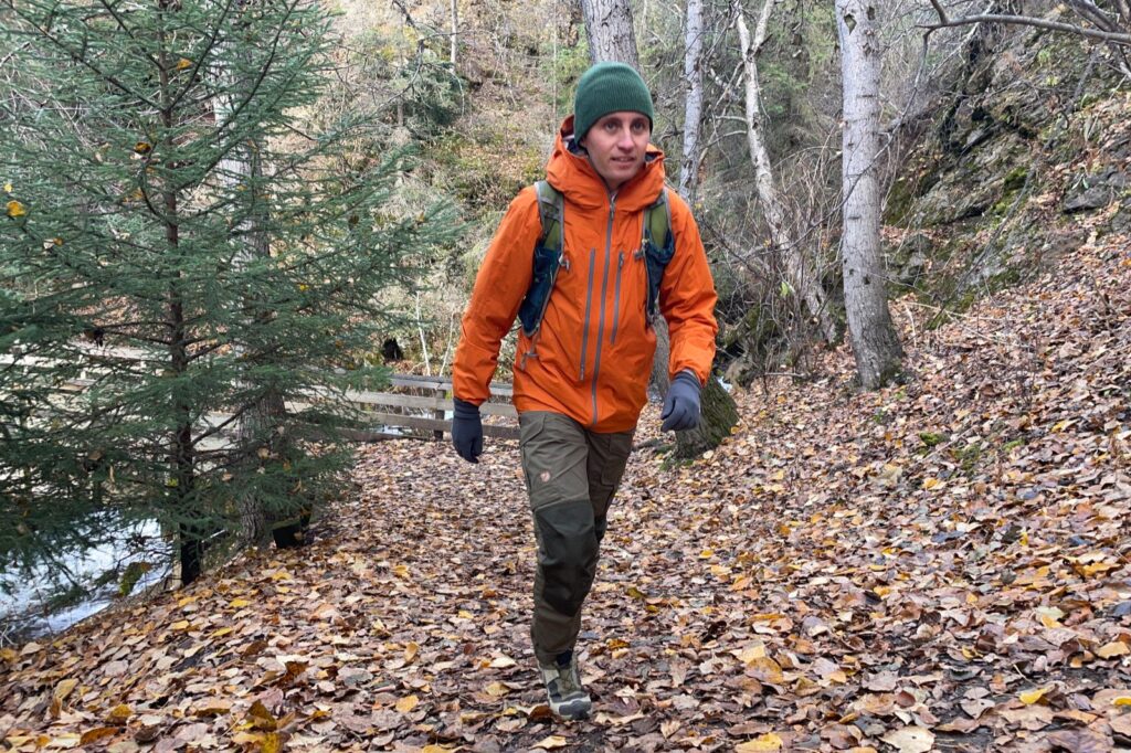 A man hikes though a forest in fall wearing an orange jacket.