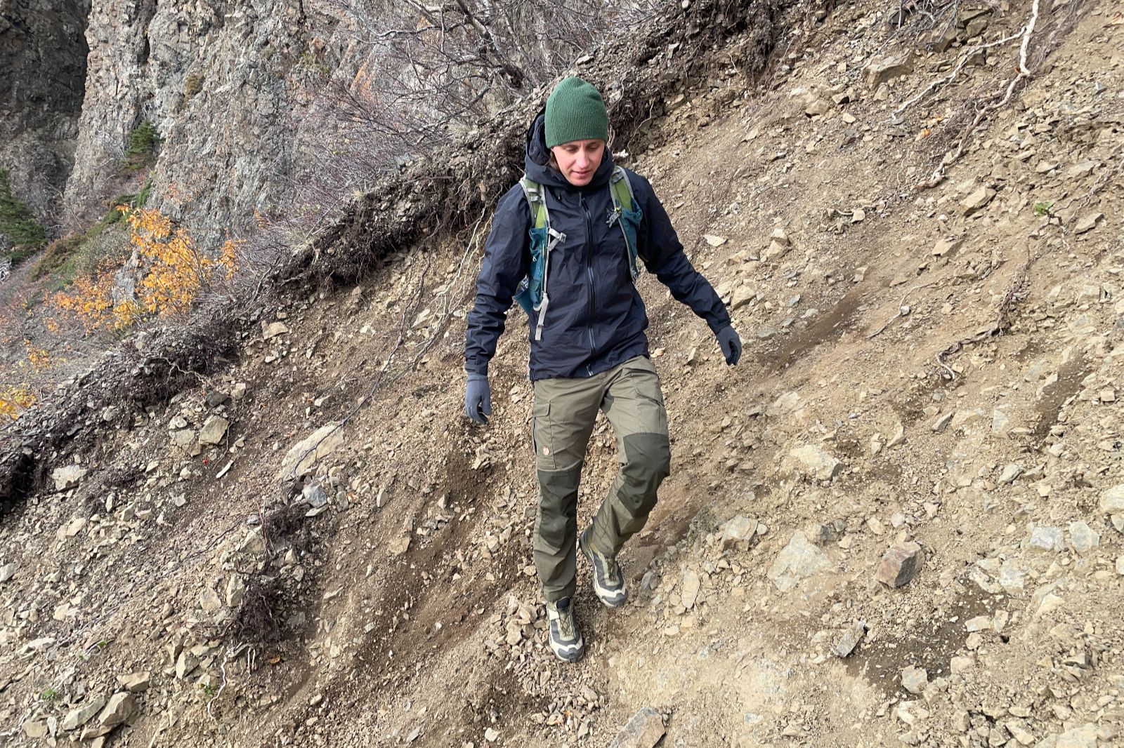A man walks across a steep slop wearing green pants and a black jacket.