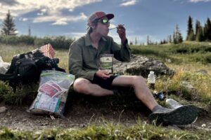a hiker sits on the ground with his gear and eats from a meal pouch. a clear bag full of food sits next to him