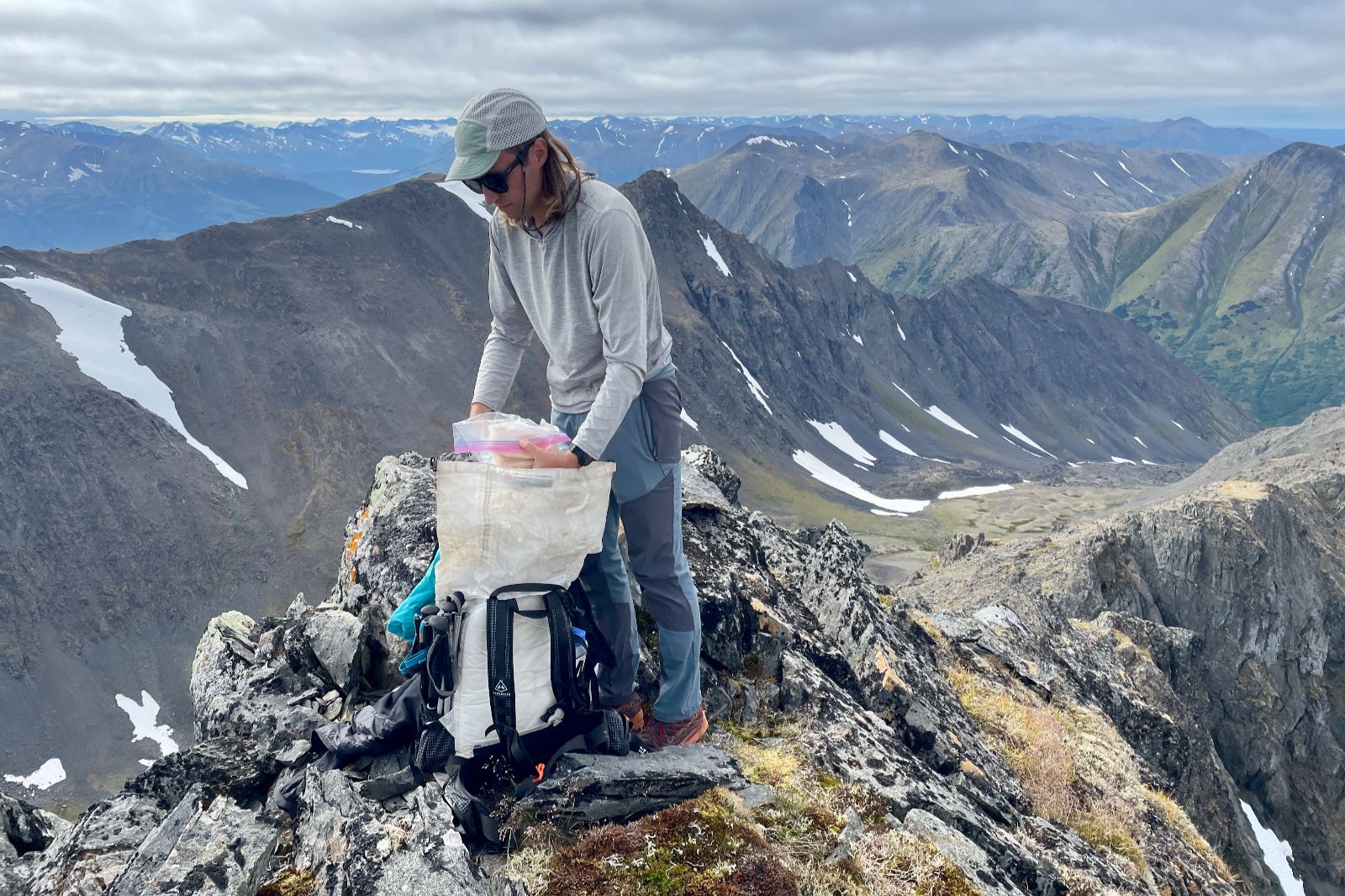 A man stands on a mountain peak putting items in his backpack.