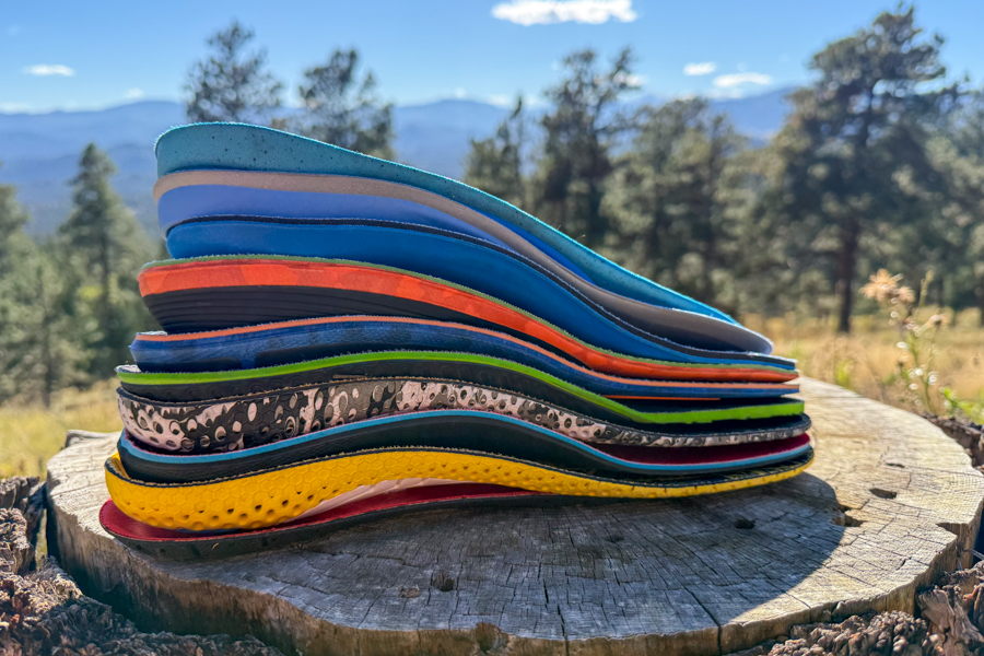 Side profile of multiple hiking insoles stacked on a tree stump, with mountains and trees in the background.