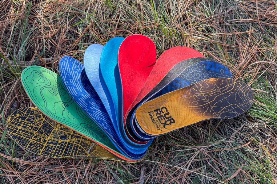 Top-down photo of multiple colorful hiking insoles fanned out on a forest floor with pine needles.