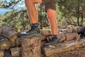 Wide shot of both feet wearing the Oboz Katabatic LT Mid boots, posed on logs with a forested mountain backdrop in golden hour lighting.