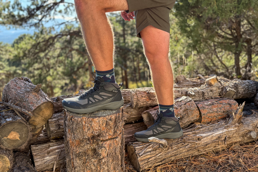 Wide shot of both feet wearing the Oboz Katabatic LT Mid boots, posed on logs with a forested mountain backdrop in golden hour lighting.