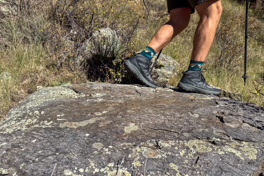 Side view of a hiker walking on a rock slab wearing Oboz Katabatic LT Mid hiking boots and teal patterned socks in a dry grassy landscape.