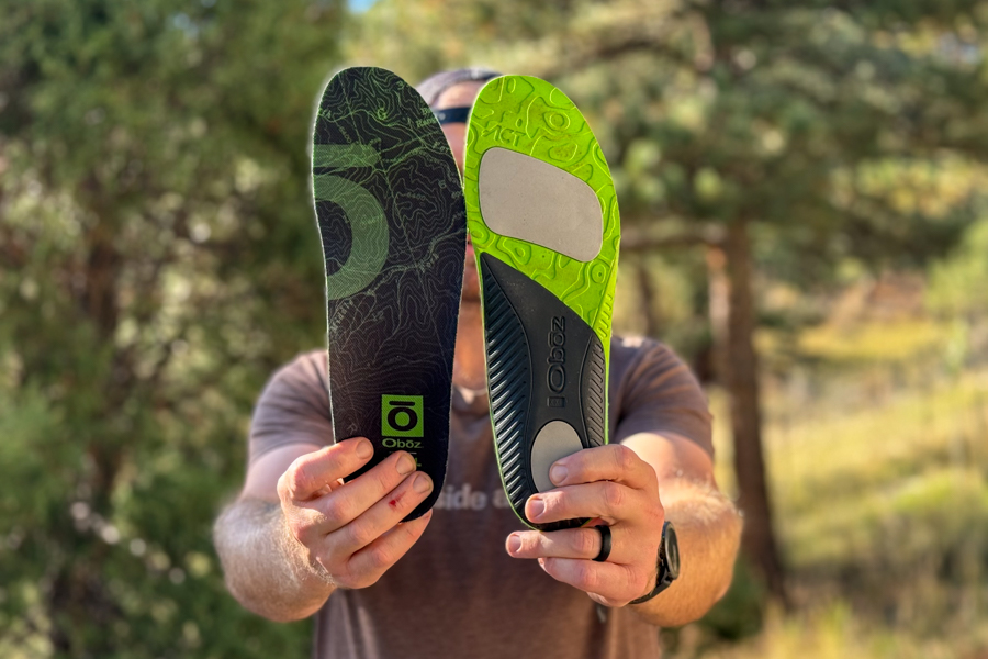 A person holds an Oboz Trail Insole in each hand, showing the top view with the logo and the bottom view with green and gray impact zones.