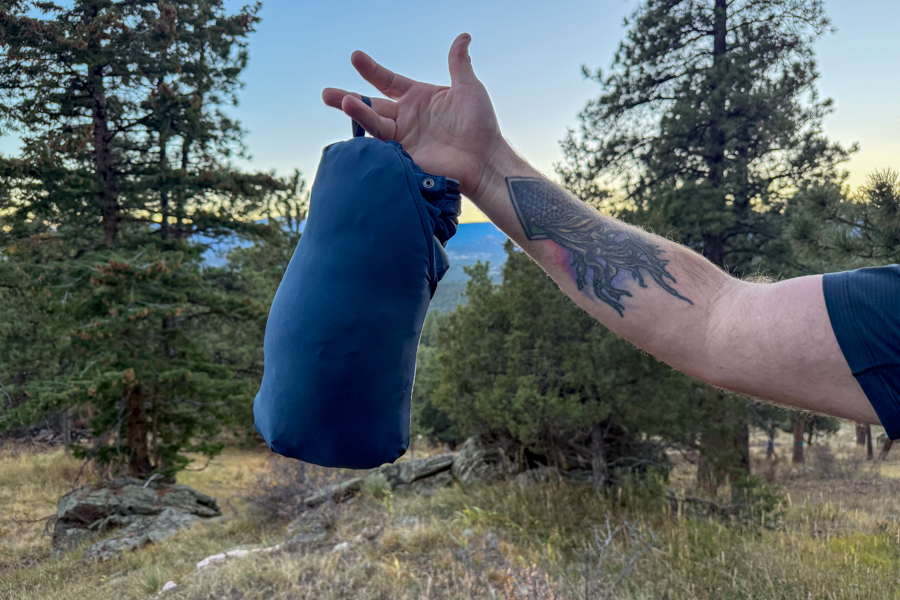 The Rab Neutrino Pro jacket fully packed into its own stuff sack, hanging from a hand against a forested mountain backdrop.
