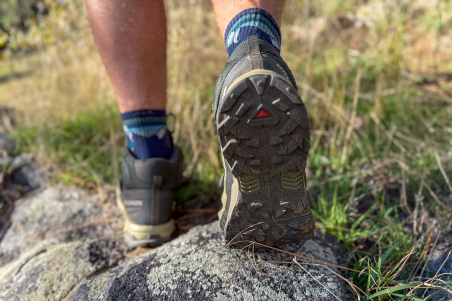 A hiker climbs a rock with the Salomon X Ultra 5, showing the aggressive Contagrip outsole tread pattern and red triangle branding designed for traction on rugged terrain.