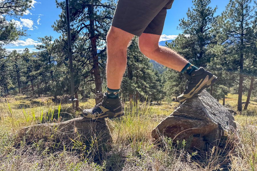 Close-up of a hiker stepping between two boulders on a sunny day, wearing mid-height Salomon hiking boots and dark shorts, with a backdrop of pine trees and a scenic mountain landscape.