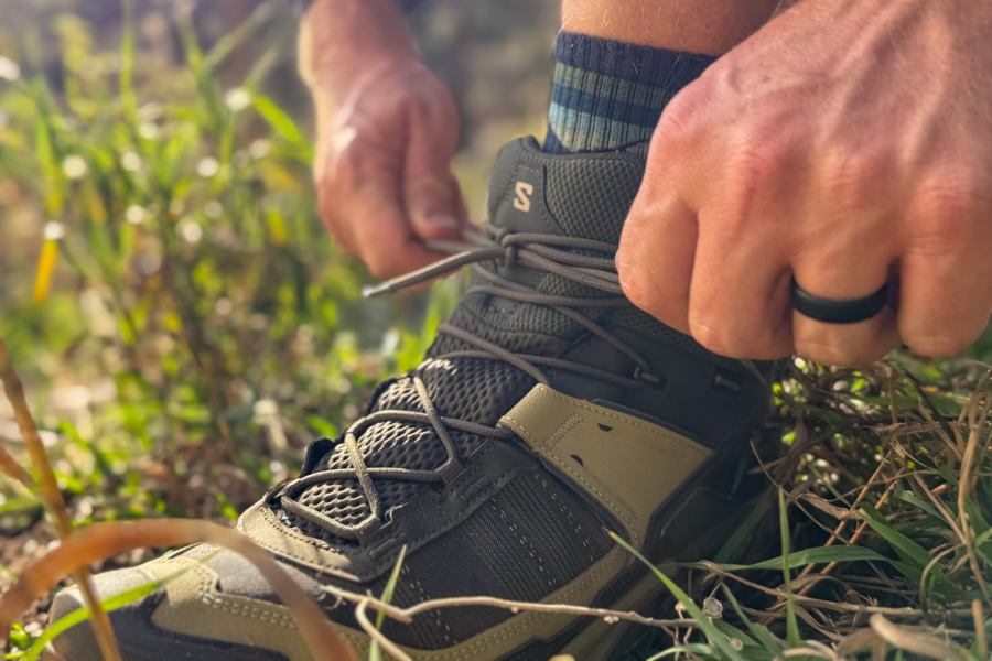 A hiker’s hands cinch down the laces of the Salomon X Ultra 5 Mid, showing the snug fit and supportive ankle collar in an outdoor grassy setting.