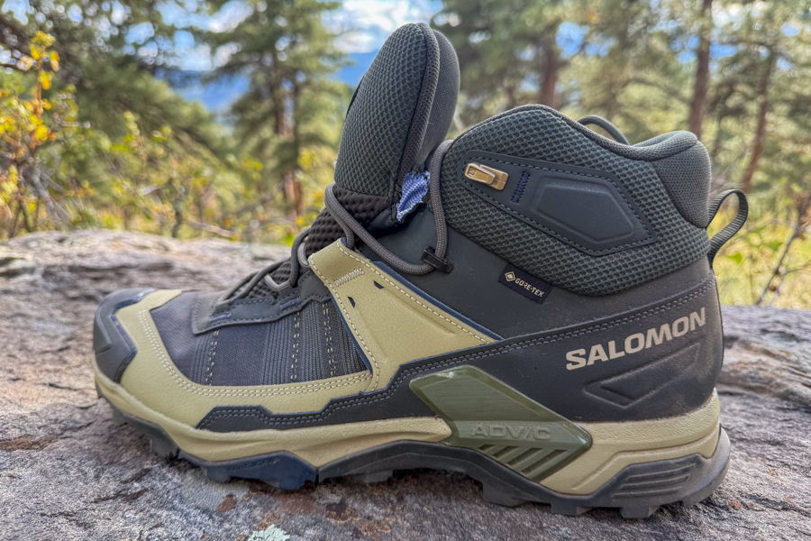 A side profile of the Salomon X Ultra 5 Mid GTX boot resting on a rock in a forested setting, with Gore-Tex branding and ADV-C Chassis visible near the heel.