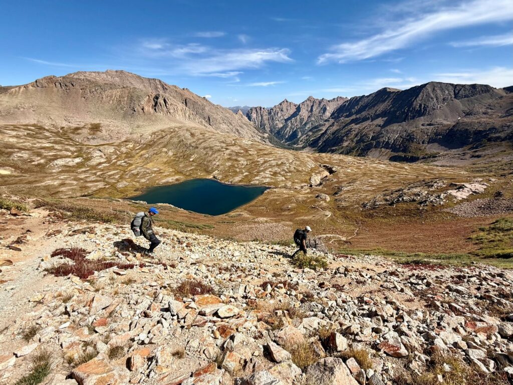 Two male backpackers descend a rocky trail that leads to a sub-alpine bowl with a small lake and short vegetation