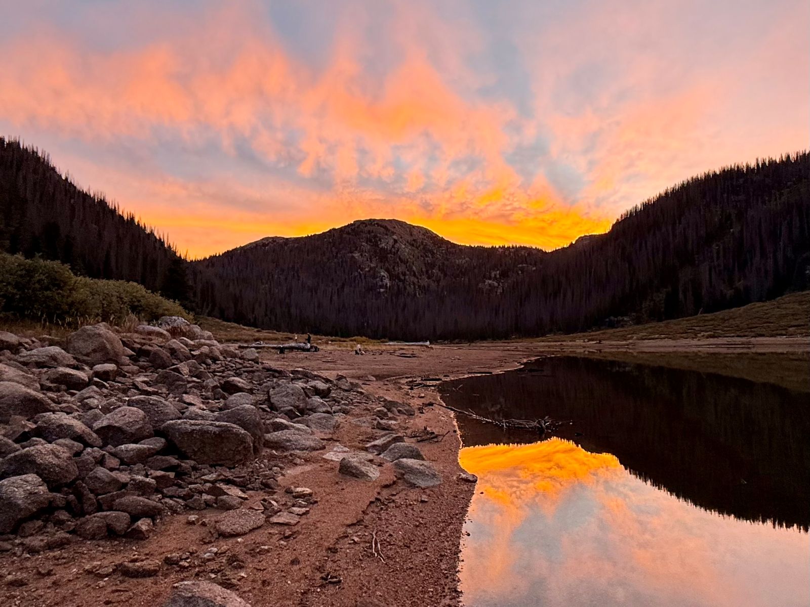 Clouds glow orange at sunset and reflect on the water of a mountain lake next to a campsite