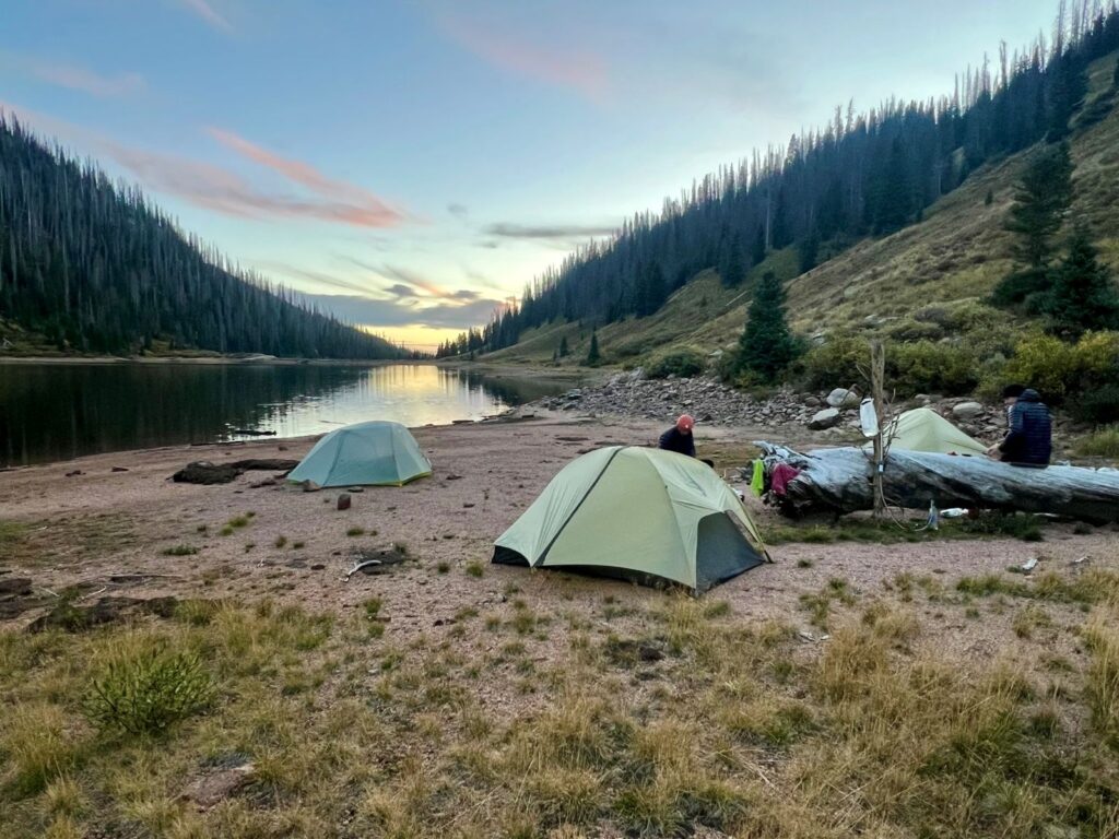 Three tents set up along a mountain lake as the sun sets behind the pines on the horizon