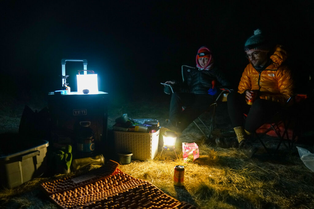 Two people sit on camp chairs in the dark next to a cooler and a Thermarest sleeping pad. The LuminAid Packlite Max 2-in-1 lantern sits on top of the cooler and the GoalZero Lighthouse 600 lantern sits on the ground, illuminating the darkness.