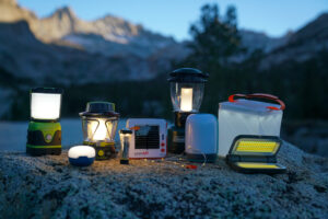 Nine lanterns- the LE 1000LM LED lantern, Black Diamond Moji, GoalZero Lighthouse 600, GoalZero Lighthouse Micro Flash, LuminAid String Light, Coleman Rugged Rechargeable, BioLite AlpenGlow 500, LuminAid Packlite Max 2-in-1, and LightUpCamp LED Solar Powered Foldable Camping Light- sit beside each other on a rock. The last remaining daylight illuminates the peaks of mountains in the background.