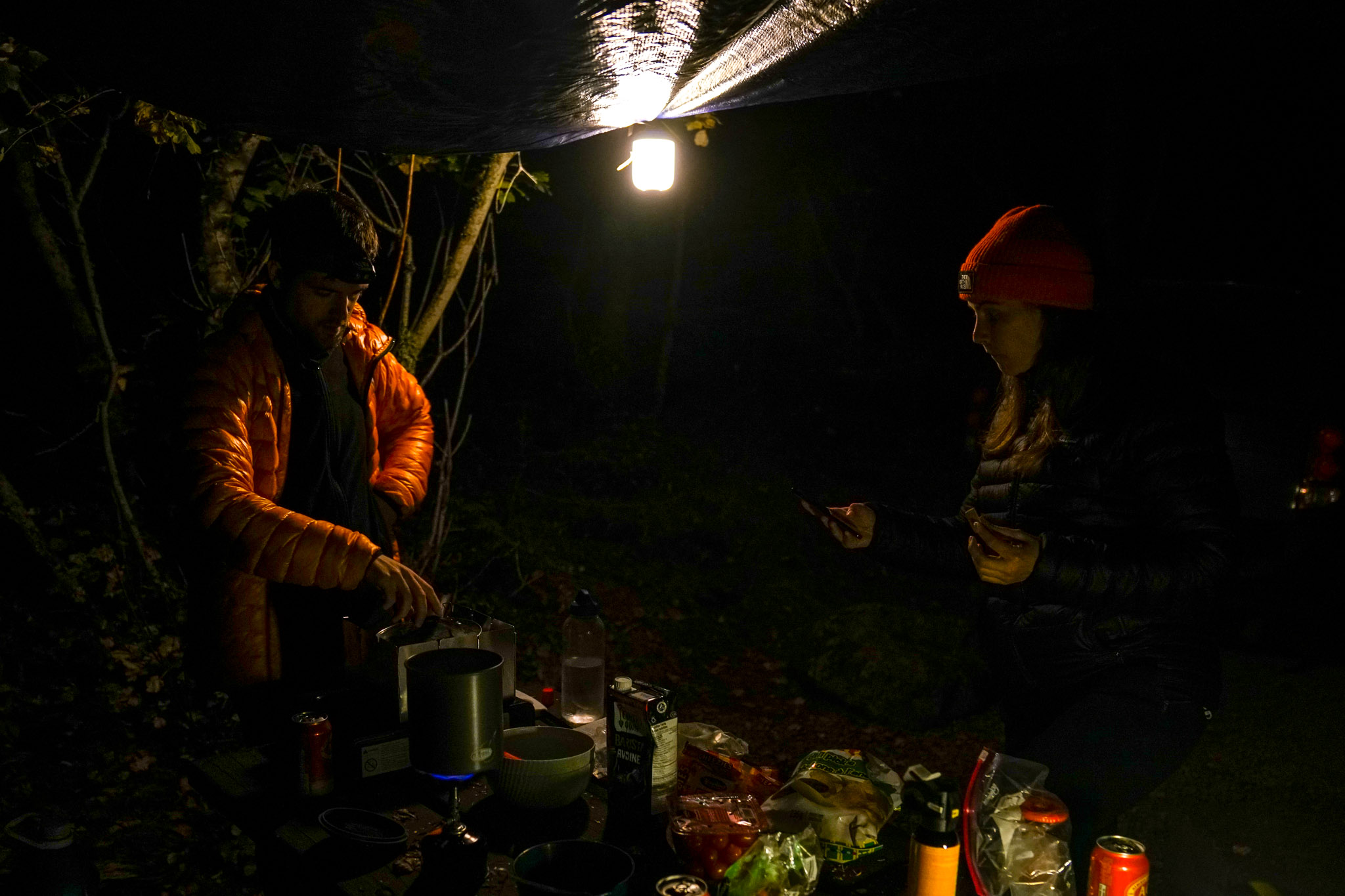 Two people stand around a picnic table covered in cooking gear in the dark. A tarp hangs above them with the BioLite AlpenGlow 500 lantern attached to its corner.