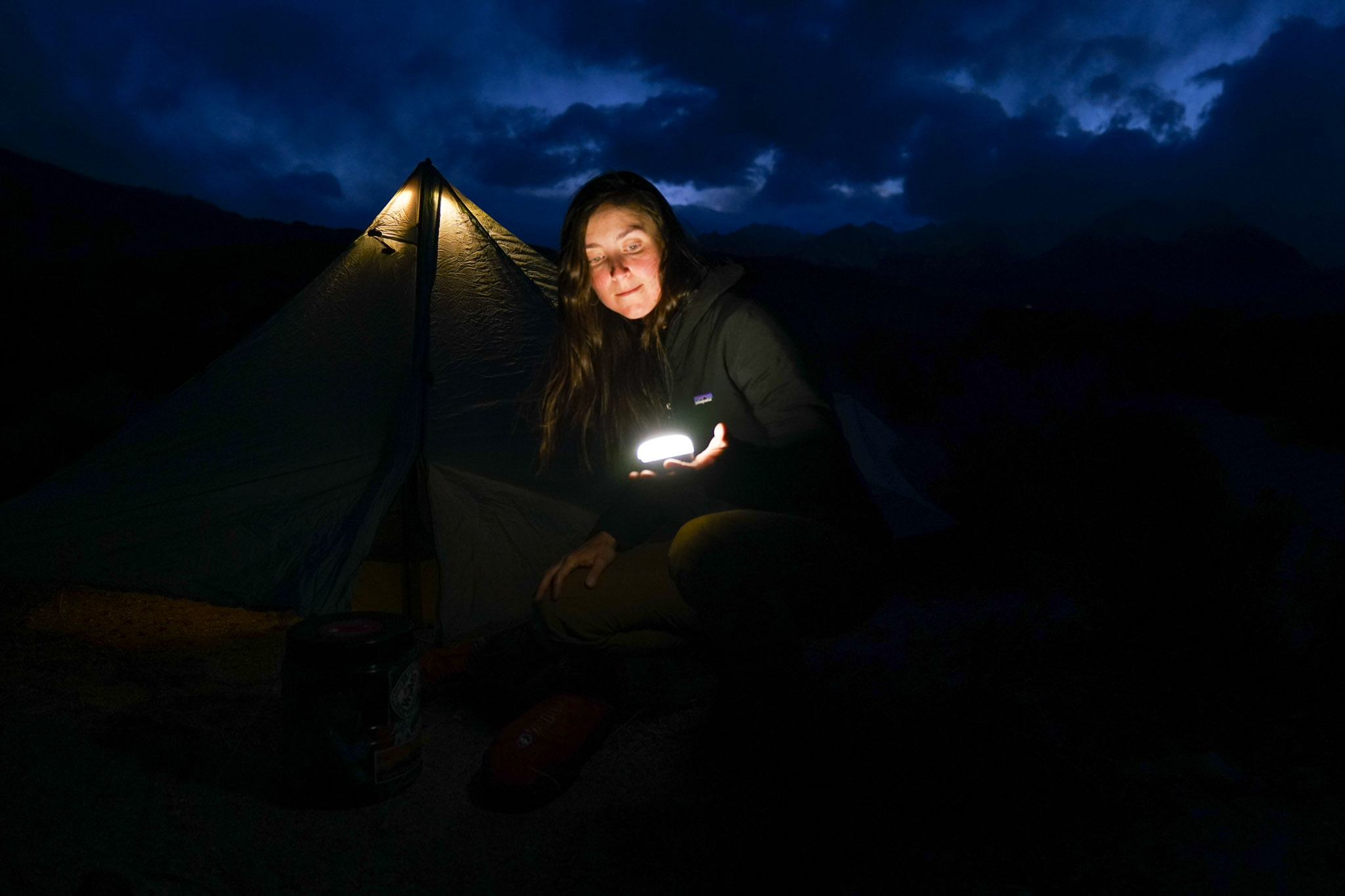 A person crouches on the ground holding the Black Diamond Moji lantern in their hand. In the background is an illuminated tent and cloudy mountaintops.