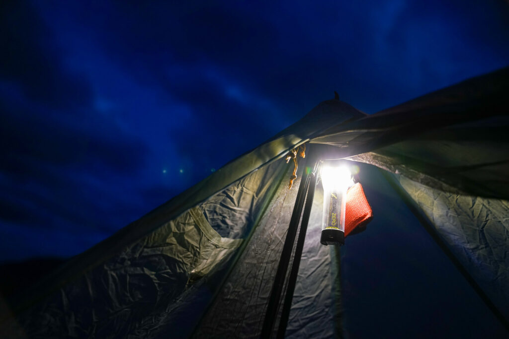 A close up looking up at the pointed top of a tent. A lit GoalZero Lighthouse Micro Flash lantern hangs at the top of the tent amidst a dark background.