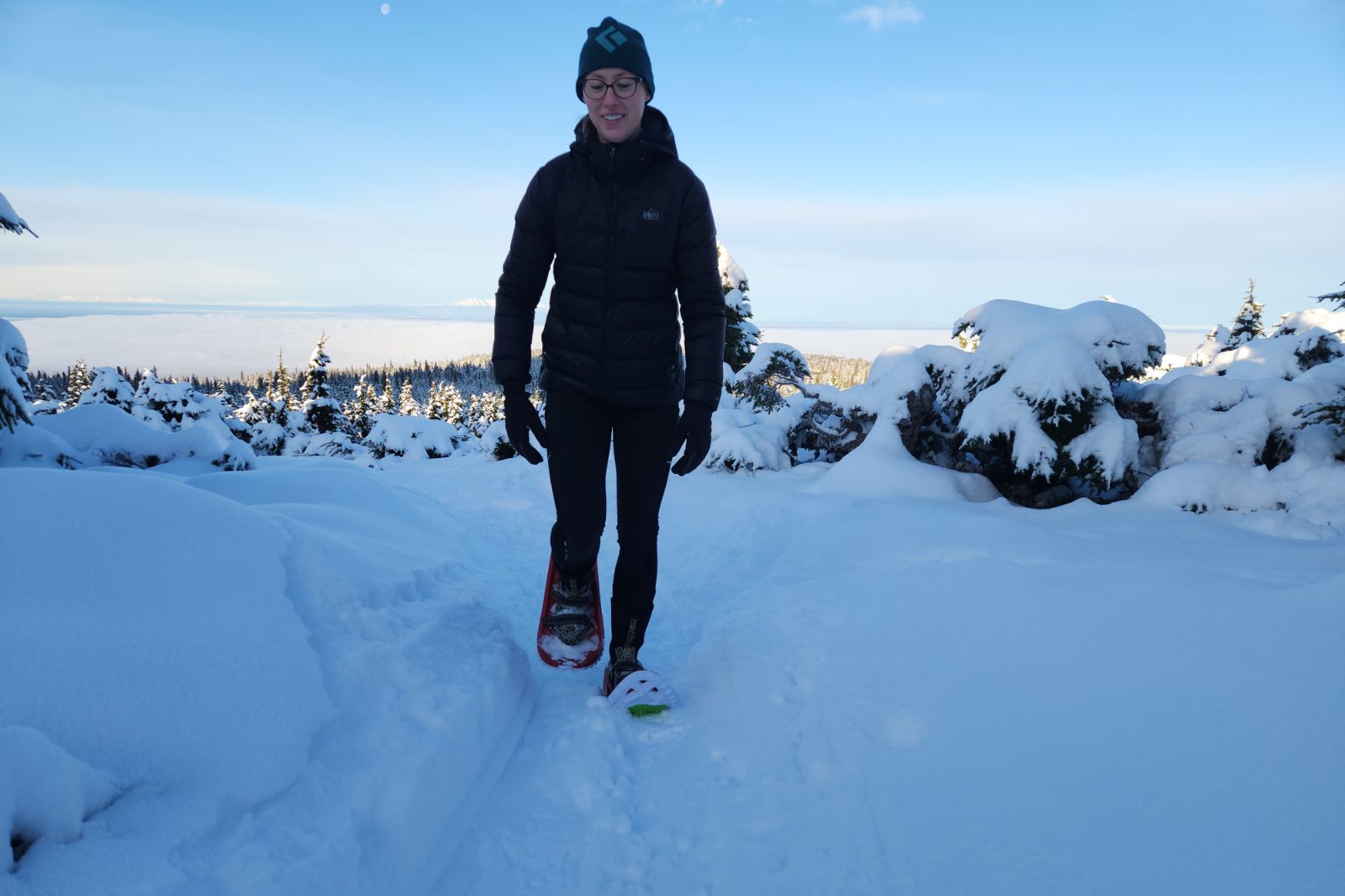 A woman walks up a snowy trail in the forest wearing snowshoes.