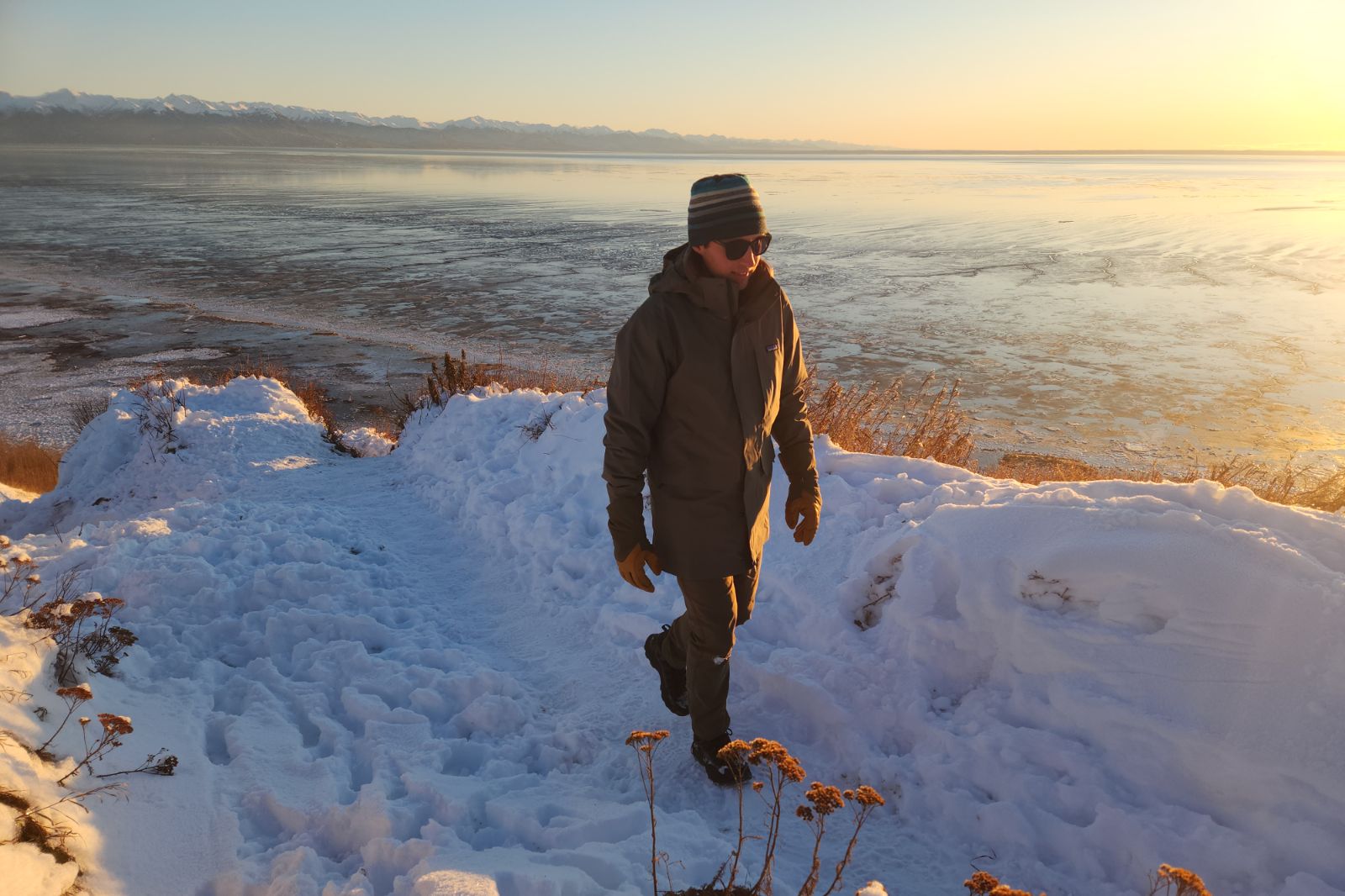 A man hikes on a snowy bluff above the ocean at sunset.