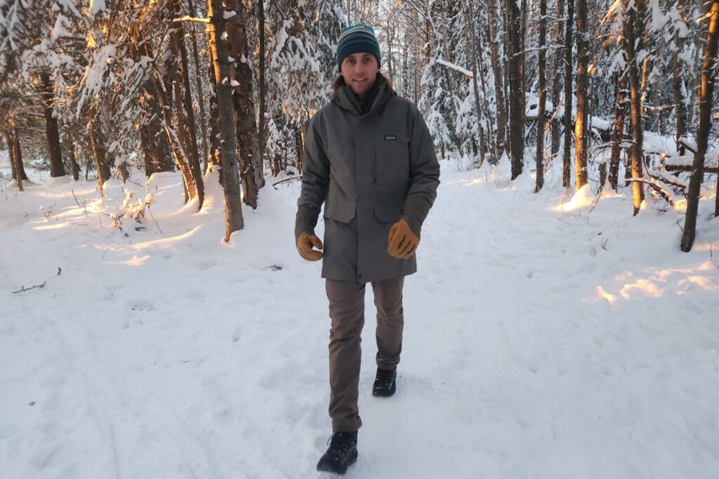 A man walks while wearing leather gloves and a green jacket in a wintry forest.