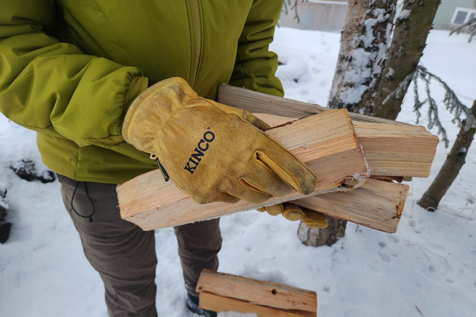 A person wearing a green jacket and yellow gloves is holding several pieces of firewood in a snowy winter setting.