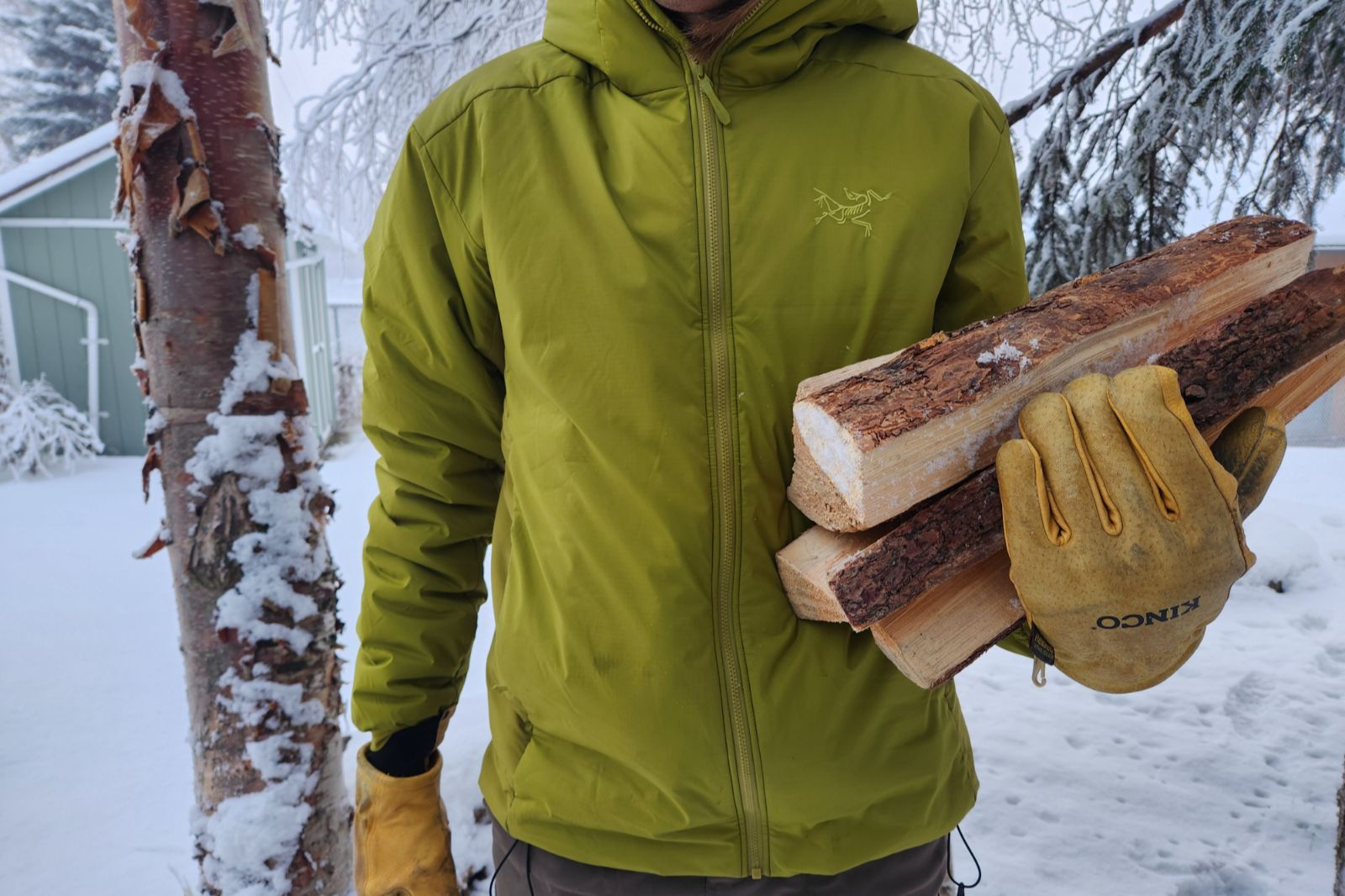 A person in a green jacket and yellow gloves stands in a snowy yard holding firewood, surrounded by snow-covered trees.