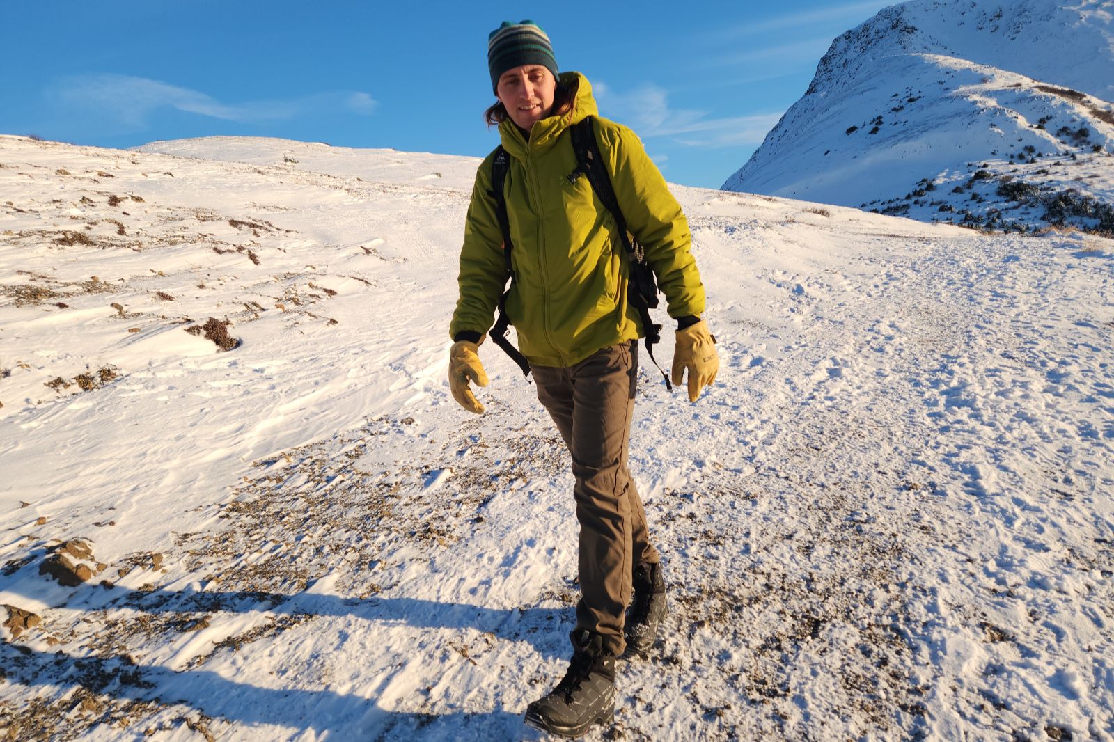 A man hikes along a snowy alpine trail in the sun.