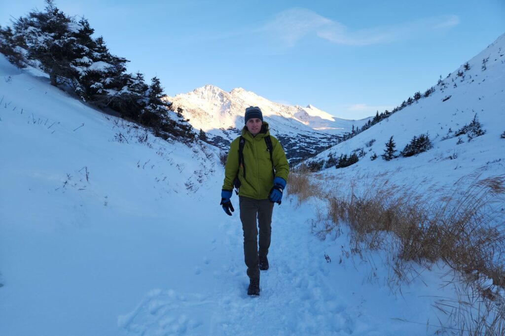A man hikes through a valley wearing a green jacket and blue gloves.