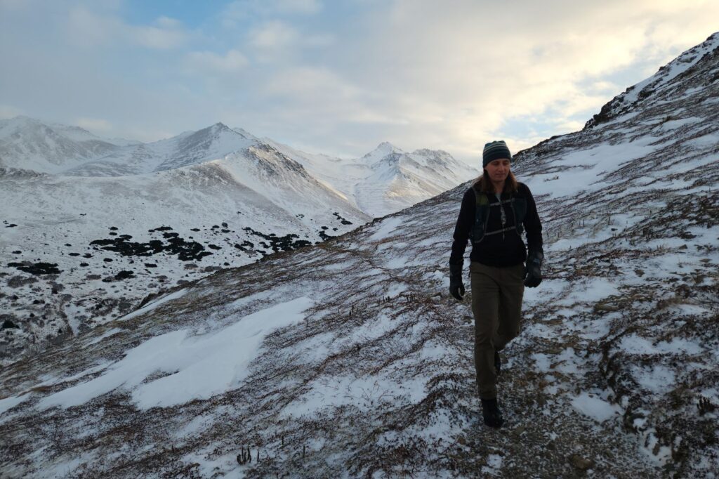 A man walks on snowy tundra with mountains behind wearing black rubber gloves.