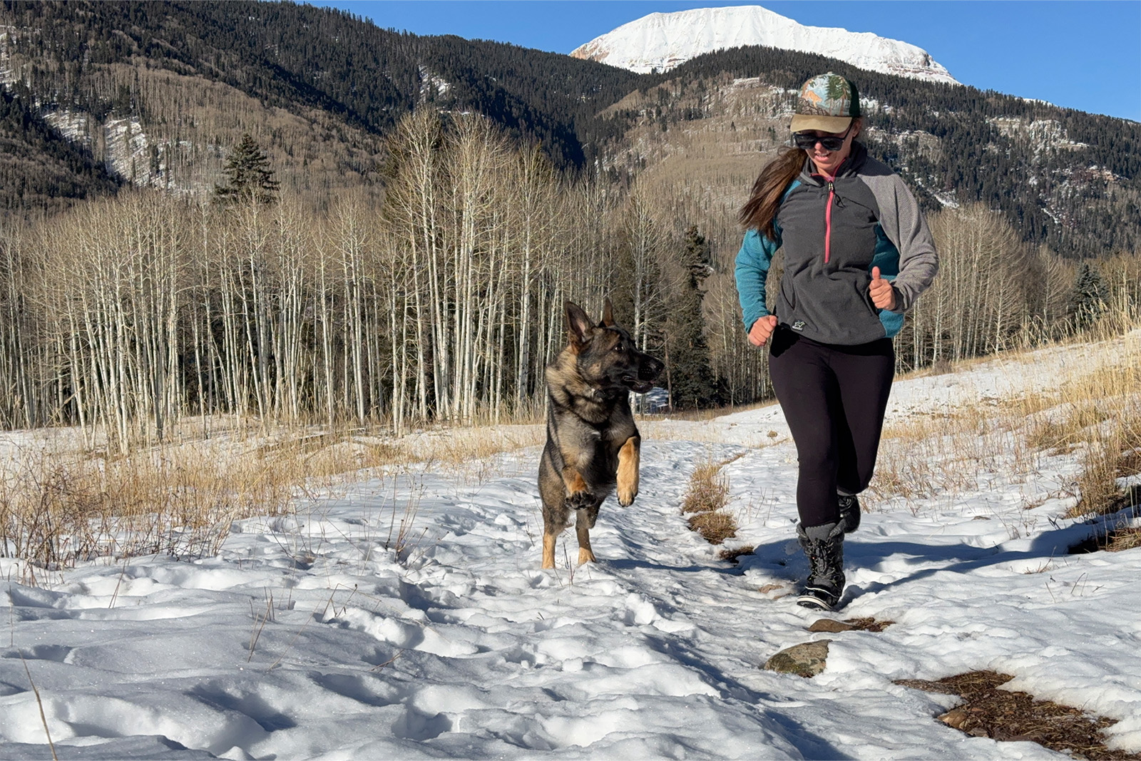 A person wearing the Dream Pairs boots is with a German Shepherd Dog up a snowy trail in a meadow. There are mountains in the background.
