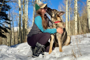 A person wearing the Dream Pair boots is petting a German Shepherd Dog in the snow. They are surrounded by aspen forest.