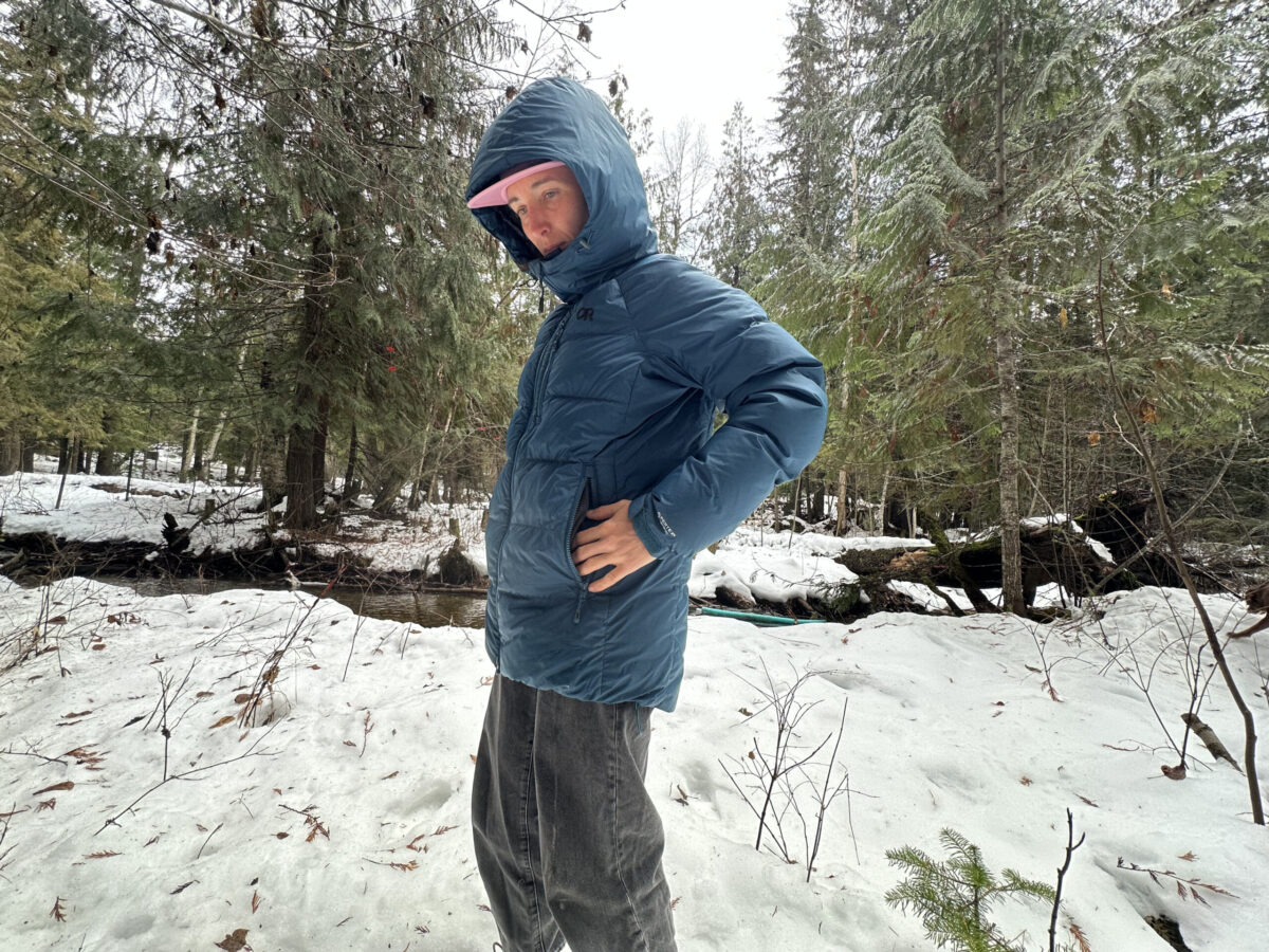 woman wearing a down jacket in the winter next to a small snowy creek and putting her hands in the pockets