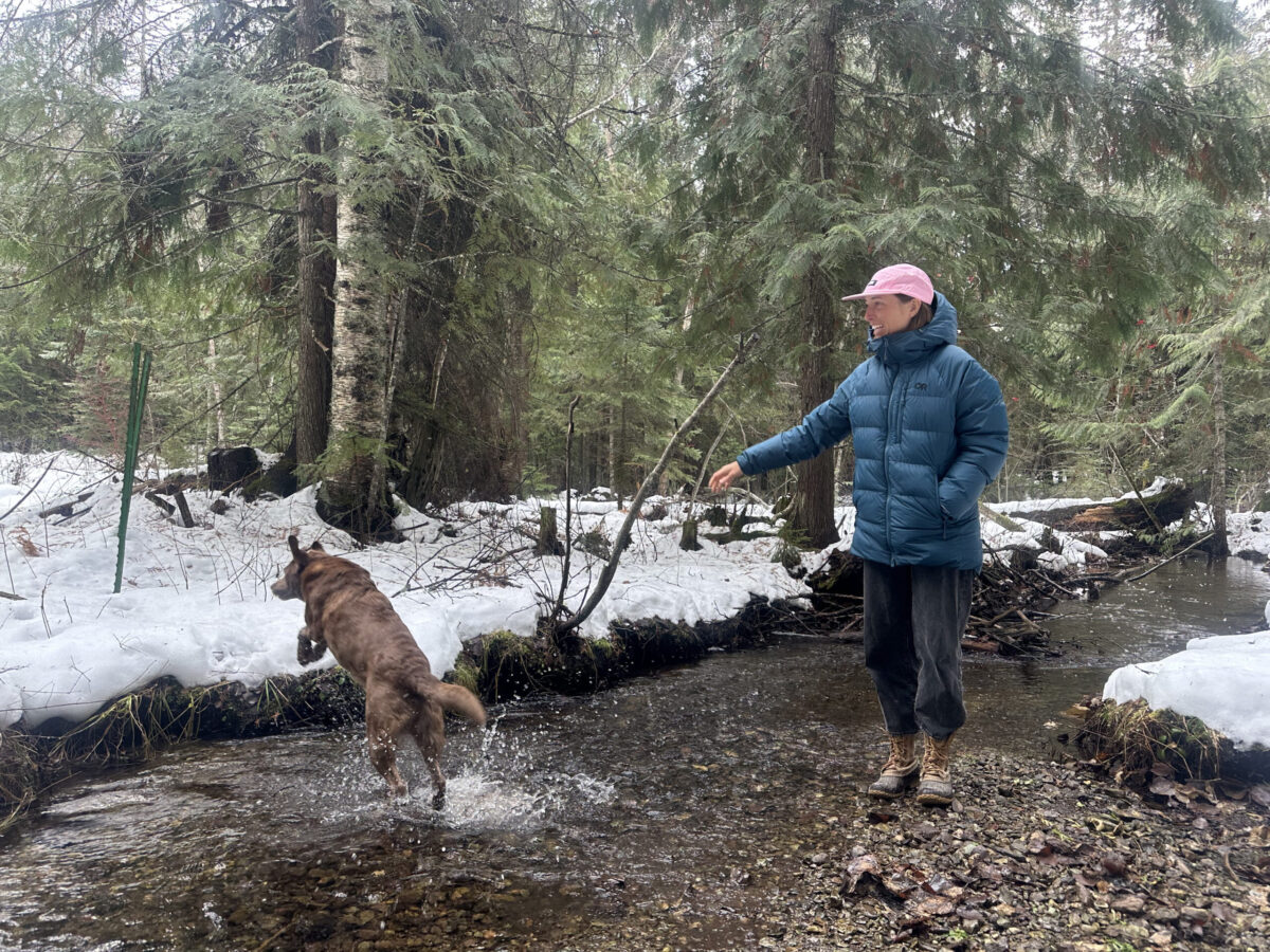 women smiles as her dog chases after a stick in a snowy forest next to a small creek