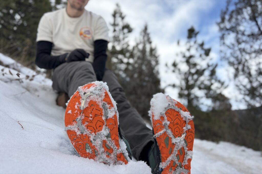 a hiker sits on a rock and shoes the bottom of his shoes with an ice cleats attached