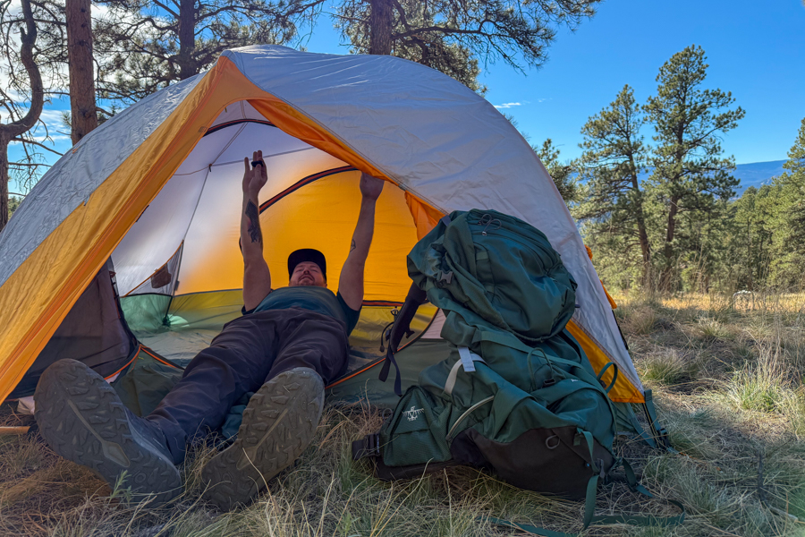 A person lying on their back inside the Mountain Hardwear Mineral King 2 All Season tent, reaching up toward the ceiling, with a large backpack resting outside the open door.
