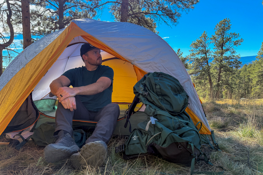 A person sitting in the open doorway of the Mineral King 2 All Season tent, looking off to the side, with a large green backpack beside the entrance in a forest meadow.