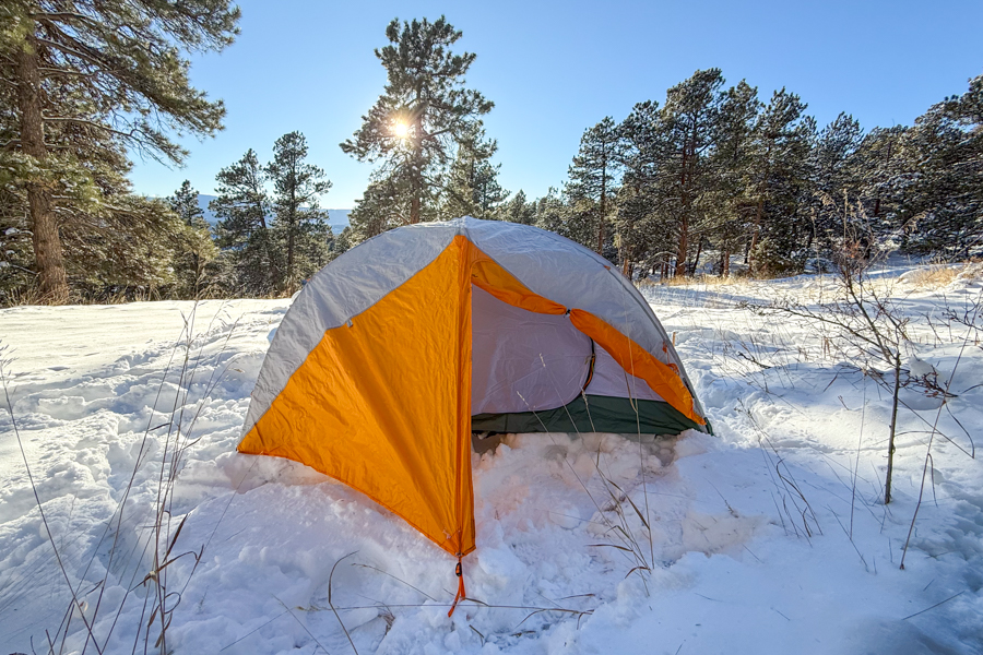 The Mineral King 2 All Season tent pitched in deep snow with its door open, illuminated by low morning sunlight filtering through surrounding pine trees.
