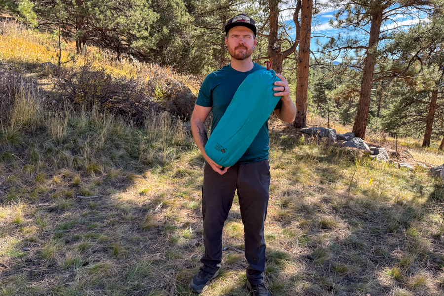 A person standing in a sunny forest meadow holding the packed Mountain Hardwear Trango 2 tent in a teal stuff sack.