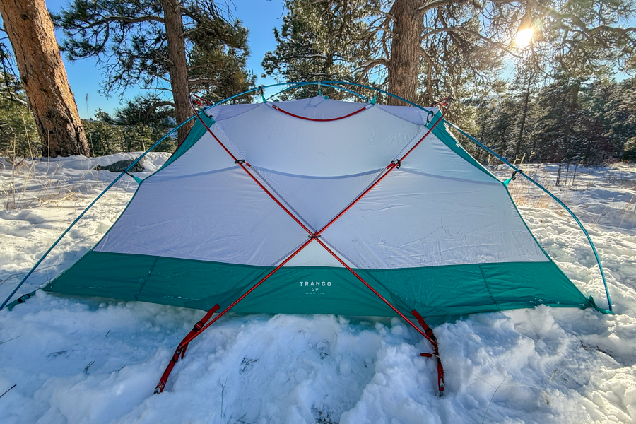 The Mountain Hardwear Trango 2 tent fully pitched on a snowy forest floor, its white and teal body and red poles illuminated by bright morning sun.